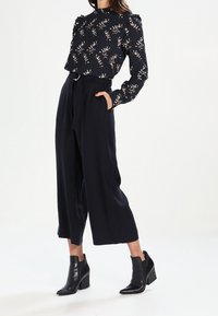 Žena v černé květinové blůze s dlouhým rukávem, s vysokým pasem a širokými černými kalhotami, a s černými podpatkovými kotníkovými botami, stojí s jednou rukou v kapse.