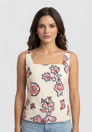 Mujer con cabello oscuro y ondulado lleva una blusa sin mangas color crema con estampado floral rosa y rojo y vaqueros azul claro sobre un fondo claro y liso.