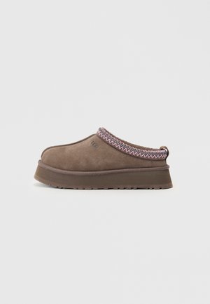 Brown suede slip-on shoe with thick sole, pink and brown zigzag stitching around collar, and small "UGG" logo on side.