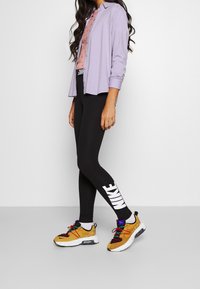 Fioletowa koszula z guzikami noszona na różowej wzorzystej bluzce, w połączeniu z czarnymi legginsami z białym logo "Nike" i żółtymi sneakersami z kolorowymi akcentami.