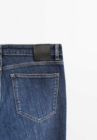Mörkblå denimjeans med en svart läderetikett vid midjan, med en enda bakficka och subtila sömdetaljer.