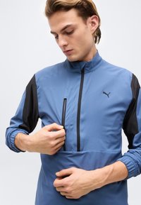 Νέος άνδρας που ρυθμίζει το φερμουάρ σε μπλε και μαύρο αθλητικό μπουφάν Puma με ψηλό γιακά, σε απλό φόντο.