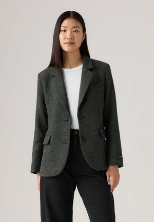 Donkergrijze blazer van gestructureerde stof, met een geknipte revers, twee knopen en voorzakken. Gekocht over een witte top, met zwarte broek.