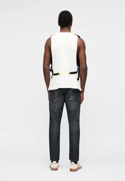 Homme portant un débardeur blanc sans manches avec des détails de sangles jaunes et noires, un jean foncé usé et des baskets blanches, debout de dos.