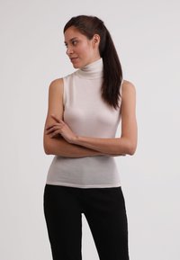 Bezrukávový top s rolákem v měkké, světlé béžové látce s hladkým povrchem, spárovaný s černými upnutými kalhotami. Modelka pózuje se zkříženýma rukama.