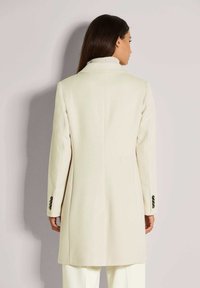 Vrouw met lang donker haar draagt een crème-kleurige op maat gemaakte jas en lichte broeken, met haar rug naar een lichtgrijze muur gekeerd.