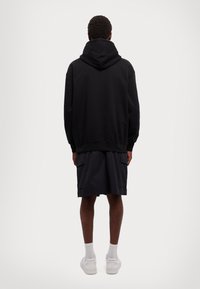 Black hooded sweatshirt with a loose fit, paired with dark shorts; model facing away, showing ribbed cuffs and hem detail.