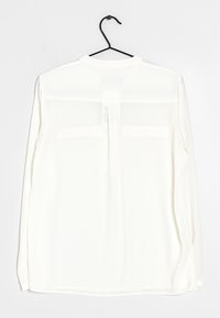Weiße Bluse mit langen Ärmeln aus transparentem Stoff, Rundhalsausschnitt und dezentem Rückfalte-Detail, hängt an einem schwarzen Haken.