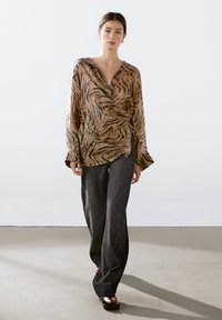 Let lys bluse i brun med tigermønster, dyb V-udskæring og lange ærmer. Kombineret med løstsiddende mørke bukser og sandaler.