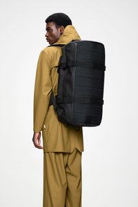 Sac à dos noir avec un devant texturé, bretelles ajustables et boucles latérales, porté sur un imperméable marron doré, design minimaliste.