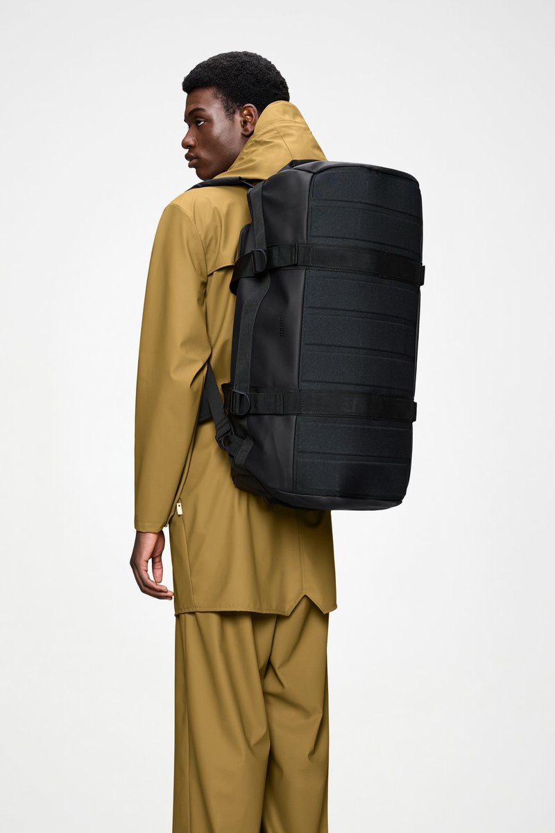 Sac à dos noir avec un devant texturé, bretelles ajustables et boucles latérales, porté sur un imperméable marron doré, design minimaliste.