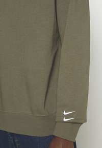 Oliveově zelená mikina Nike s texturovaným povrchem, se širokým rukávem s dvěma bílými akcenty loga. Uvolněný střih a zesílené švy.