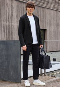 Blazer negro con dos bolsillos frontales sobre una camiseta blanca, combinado con pantalones negros y zapatillas blancas. Lleva un bolso de cuero negro.