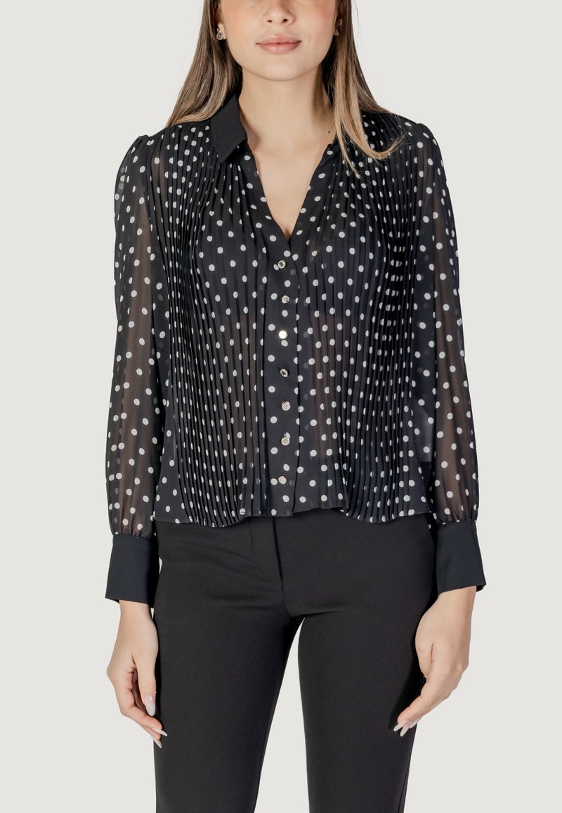 Blusa nera trasparente con pois bianchi, pieghe verticali, chiusura con bottoni e polsini sartoriali. Adatta per abbigliamento elegante o casual.