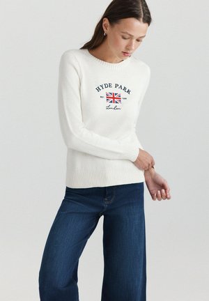 Femme portant un pull blanc à manches longues avec le texte Hyde Park London et un graphisme du drapeau britannique, associé à un jean bleu foncé, debout sur un fond blanc.