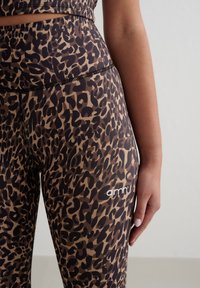 Leggings med leopardmønster i brunt og svart, laget av glatt stoff med en jevn tekstur, med en brodert logo på siden.