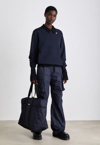 Felpa blu navy con colletto, abbinata a pantaloni cargo blu navy e con una grande borsa a tracolla. Il materiale sembra texturizzato e resistente.