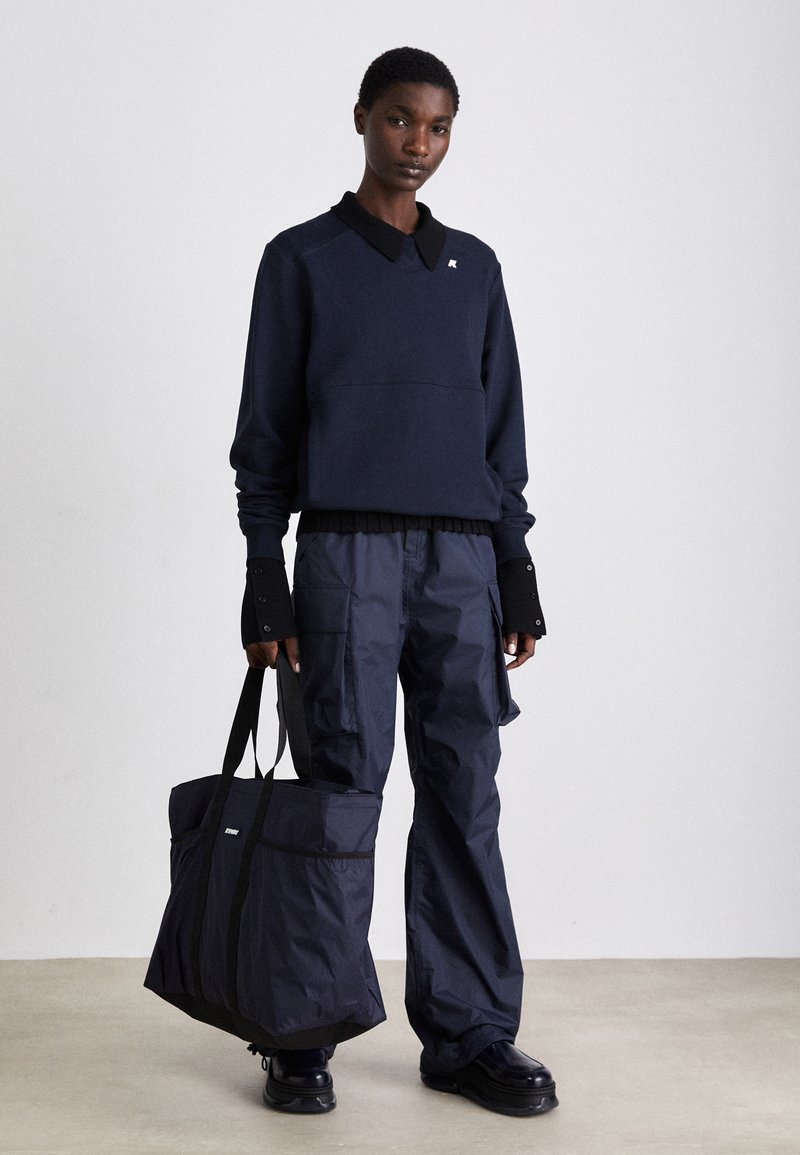 Felpa blu navy con colletto, abbinata a pantaloni cargo blu navy e con una grande borsa a tracolla. Il materiale sembra texturizzato e resistente.