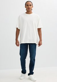 Camiseta de algodón blanca de gran tamaño, de corte relajado, combinada con unos vaqueros de denim azul oscuro. Calzado: zapatillas blancas con detalles texturizados.