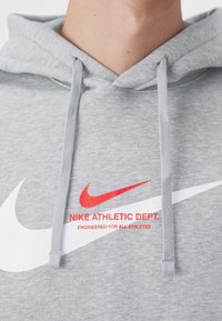 Szara bluza z kapturem z logo Nike, czerwonym napisem "NIKE ATHLETIC DEPT. ENGINEERED FOR ALL ATHLETES" oraz dwoma sznurkami widocznymi przy szyi.