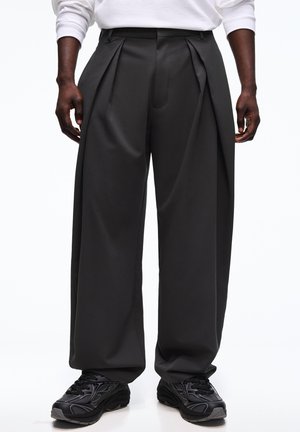 Mężczyzna w luźnych czarnych plisowanych spodniach i czarnych trampkach, stojący na białym tle, z rękami swobodnie opuszczonymi wzdłuż ciała.