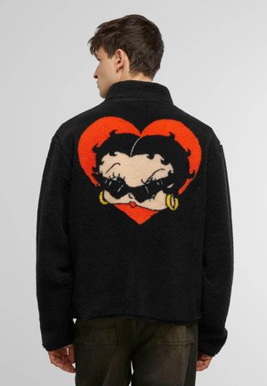 Jachetă din fleece neagră cu guler înalt, cu un design în formă de inimă roșie și un personaj de desene animate colorat pe spate, accentuată de cercei galbeni.