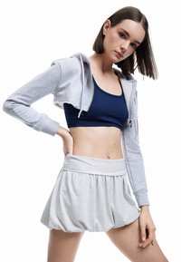 Joven mujer vestida con un top corto azul marino, una sudadera corta gris claro y una falda corta de tiro alto en gris claro, con un fondo blanco.