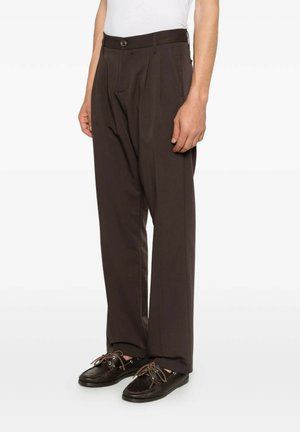 Uomo che indossa pantaloni plissettati marrone scuro e scarpe da barca in pelle nera con lacci marroni, in piedi su sfondo bianco.