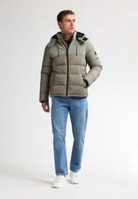 Geacă puffer în verde măsliniu cu o secțiune superioară contrastantă, închidere cu fermoar, buzunare laterale și glugă. Asortată cu blugi de culoare bleu deschis și adidași albi.