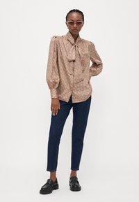 Blusa marrone a fantasia con colletto a fiocco, maniche lunghe a sbuffo, abbinata a jeans slim blu scuro e mocassini neri chunky.