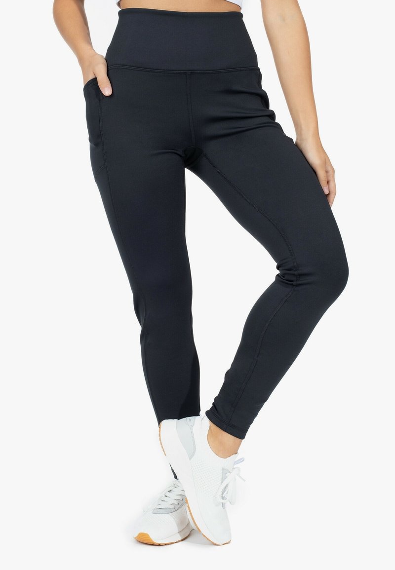 Leggings neri a vita alta con una texture liscia, dotati di tasche laterali e design aderente, abbinati a sneakers bianche con dettagli grigi.