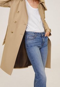 Osoba w beżowym trenczu noszącym biały top na ramiączkach, włożony w wysokie niebieskie dżinsy, z jedną ręką w kieszeni.