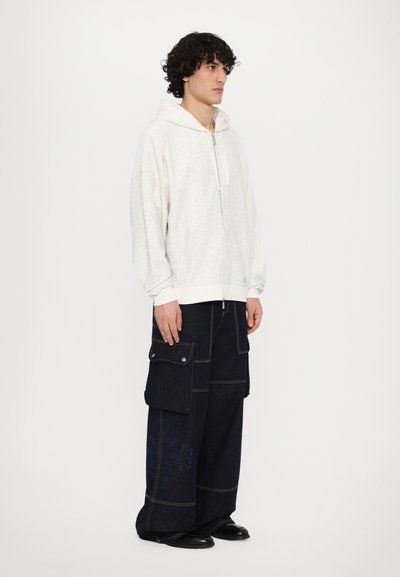 Sweat à zip blanc avec un motif texturé et de grandes poches avant, associé à un pantalon cargo en denim foncé à jambes larges, avec des poches latérales et des coutures.