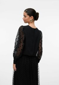 Mustaga mustriga polka-dot varrukatega must särk ja tekstureeritud kehaosaga. Omab ümmargust kaelust ja väikest tagumist sulgemise detaili. Stiliseeritud sobiva seeliku juurde.
