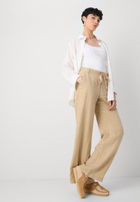 Beige linen pants with a drawstring waist, wide leg, and side pockets. Paired with a white tank top and a lightweight white shirt.