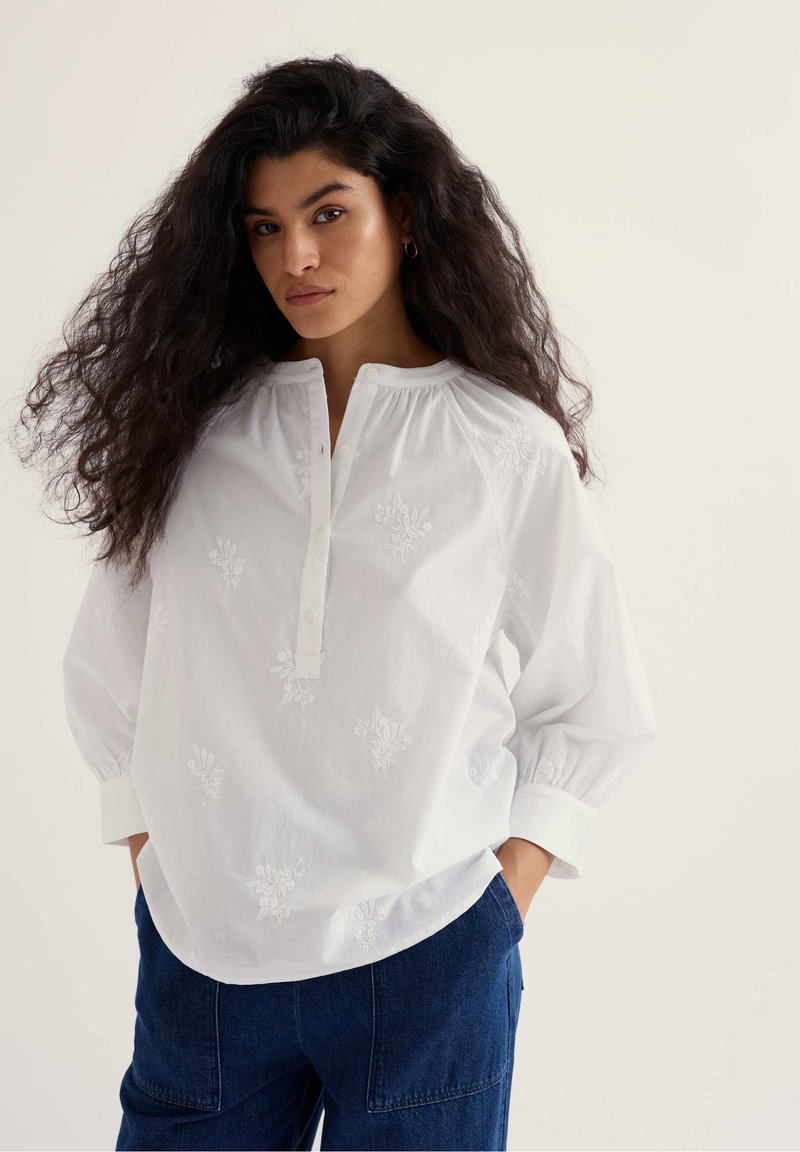Femme aux longs cheveux bouclés portant un chemisier blanc brodé avec des boutons et un pantalon bleu, debout les mains dans les poches.