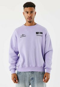 Hombre con cabello rizado corto, vistiendo un sudadera ligero de color púrpura con logos negros y jeans de color azul claro, posando contra un fondo blanco plain.