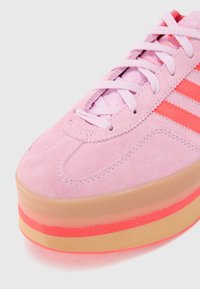 Gros plan sur une basket en suède rose avec des lacets blancs, des bandes rouges sur le côté et une épaisse semelle en gomme composée de couches rouges et beiges.