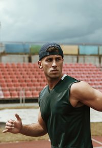 Egy férfi sportoló ujjatlan, sötétzöld sportingben és sötétkék sapkában áll, sima textúrával, egy stadion háttér előtt.