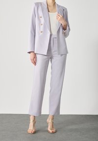 Ljus lavendel kostym med en dubbelknäppt jacka med guldknappar, skräddarsydda byxor och sandaler med guldremmar på ett grått golv.