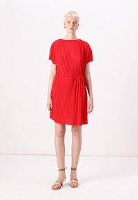 Vestido plissado vermelho com mangas curtas e cintura elástica, apresentando um corte solto. Usado com sandálias bege com tiras.