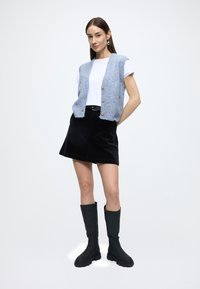 Lichtblauwe gebreide spencer met korte mouwen, knoopsluiting aan de voorkant; wit T-shirt eronder, zwarte corduroy rok, kniehoge zwarte laarzen.