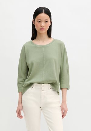 Kobieta z prostymi czarnymi włosami nosi luźny jasnizielony top z rękawami do ćwiartki oraz wysokie, kremowe spodnie na białym tle.