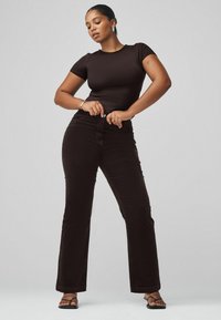 Schwarzes, eng anliegendes Kurzarm-Oberteil kombiniert mit hoch taillierten, dunkelbraunen Schlagjeans. Model trägt schwarze Sandalen und präsentiert glatte, strukturierte Stoffe.