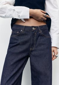 Oseba dviguje temno moder telovnik, da razkrije trebuh, oblečena v belo srajco z gumbi za manšete, temno modre kavbojke in več srebrnih prstanov na rokah.