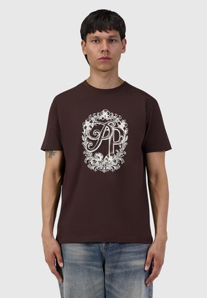 Young man with short dark hair wears a brown t-shirt with a white floral frame design and the letters P and R on the front.