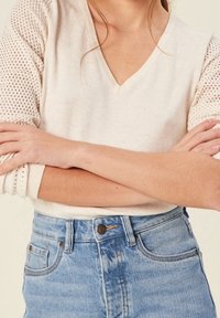 Femme les bras croisés portant un pull en maille crème avec des manches en mesh et un jean en denim bleu clair à taille haute.