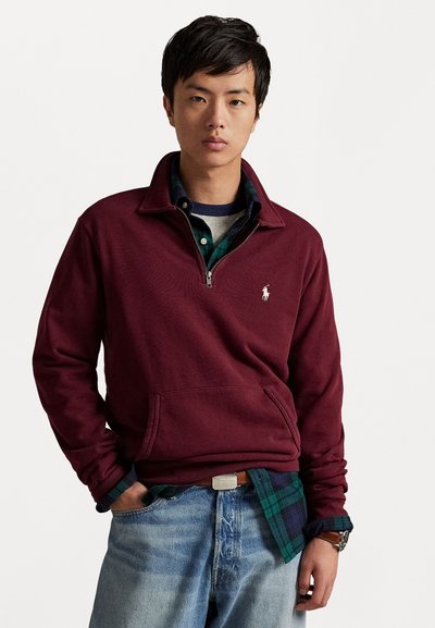Burgundero pullover aus Fleece mit halben Reißverschluss-Kragen, Vordertasche und kleinem gesticktem Logo. Getragen über einem grün karierten Hemd und hellblauem Denim.