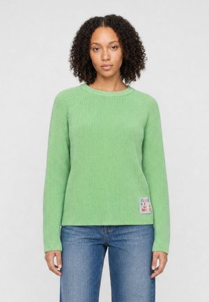 Woman with curly hair wearing a light green knit sweater and blue jeans standing against a plain background.