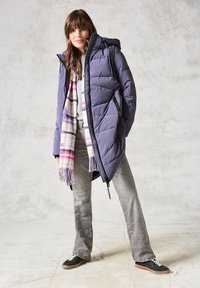 Chaqueta de plumas morada con capucha, adornada con costuras en zigzag y bolsillos con cremallera, combinada con una bufanda de cuadros y jeans grises. Zapatillas negras.